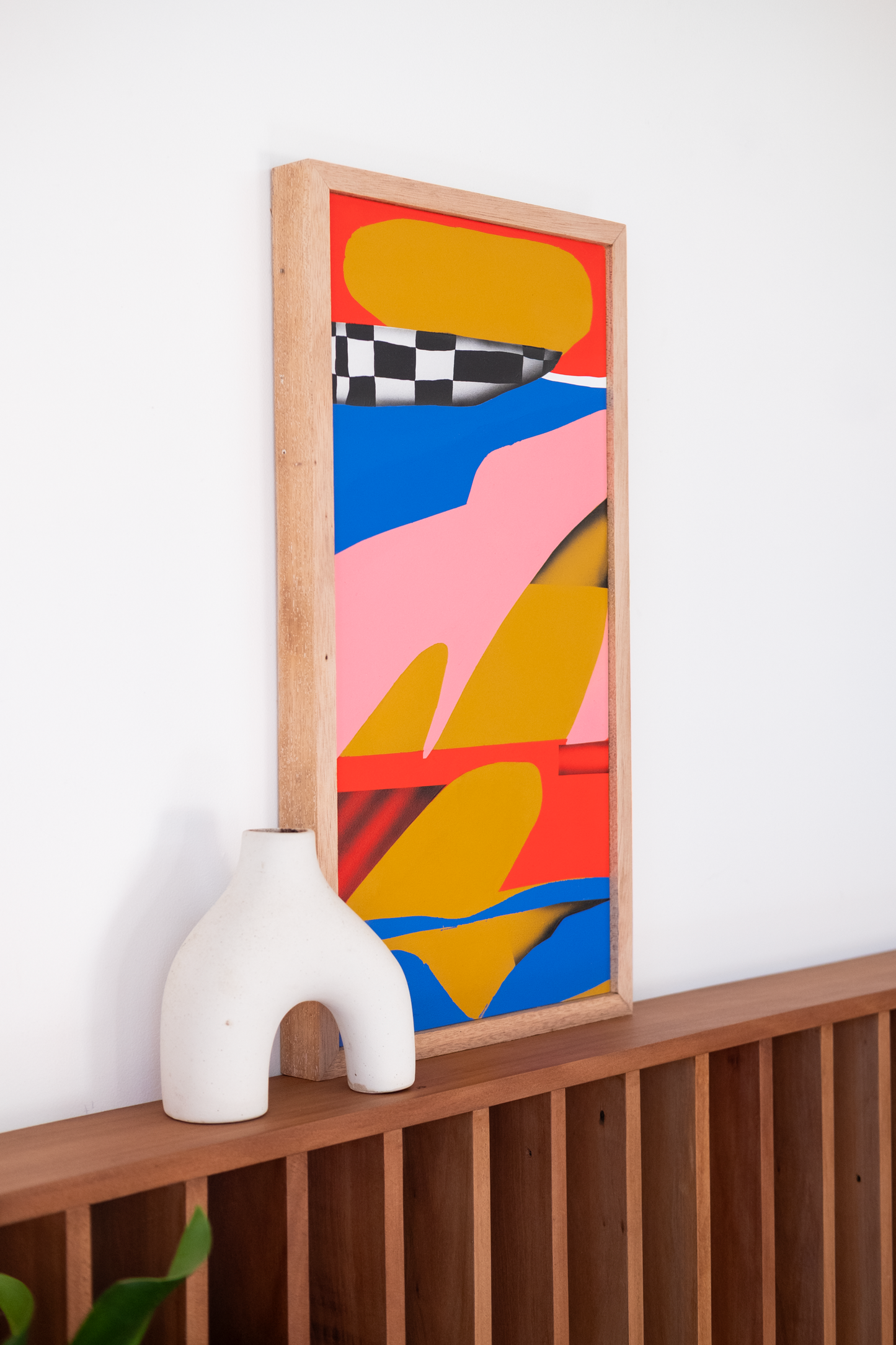 Colorful abstract painting in a wooden frame on a white wall with a white sculpture below.